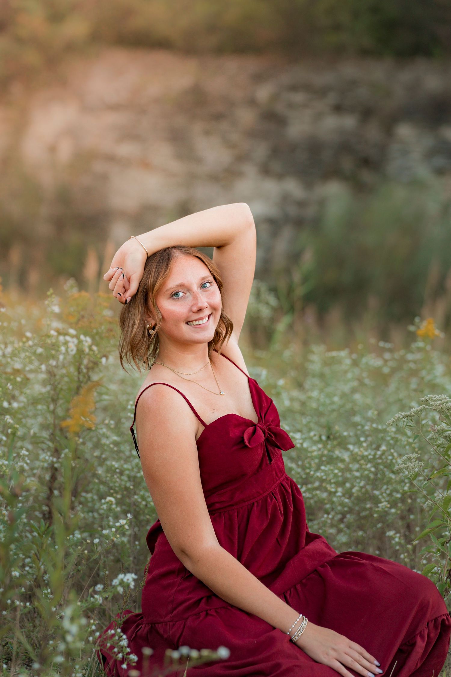 A woman in a burgundy dress poses gracefully among wildflowers in a dreamy outdoor setting during golden hour.