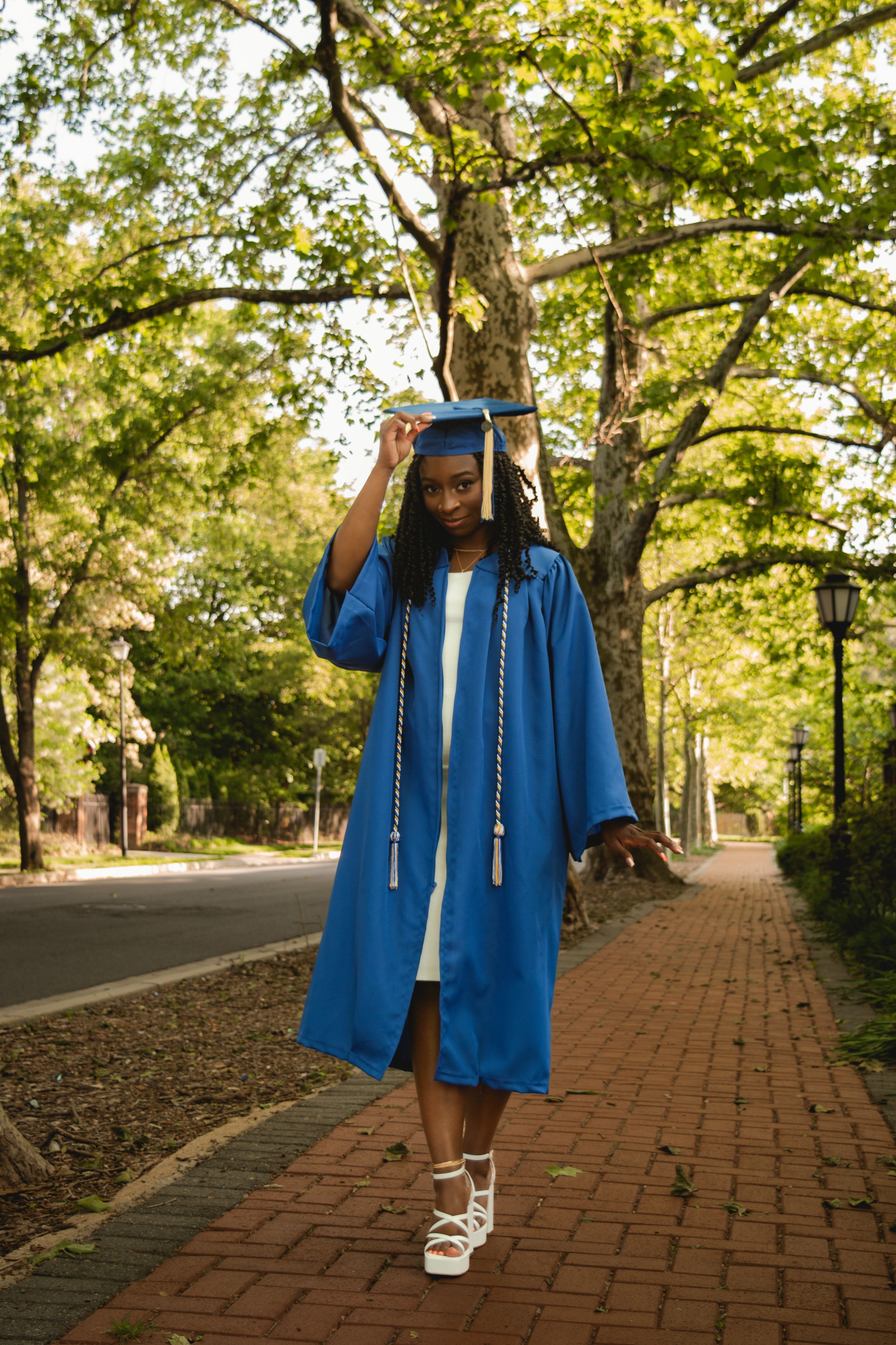 Graduate in blue cap and gown walking on a brick pathway surrounded by lush green trees on a sunny day.