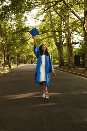 A graduate in blue cap and gown celebrates their achievement by jumping with joy in a tree-lined pathway.
