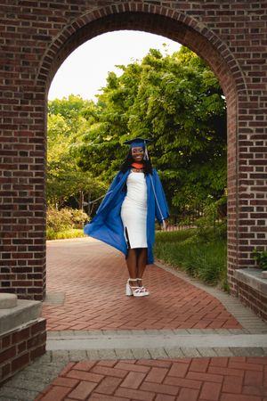 Graduate in blue cap and gown walks along a brick pathway surrounded by lush greenery on a sunny day.