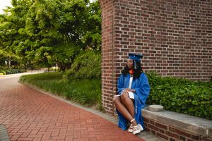 Graduate in blue cap and gown sits on brick ledge along curved brick pathway on sunny day.