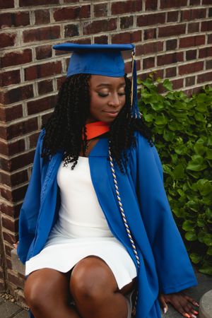 Graduate in blue academic regalia poses against a brick wall with green foliage in the background.