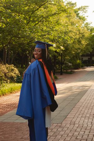 Graduate in blue academic regalia poses on tree-lined brick walkway during golden hour.