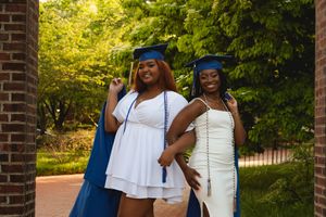 Graduates in white dresses and blue caps celebrate together outdoors on a sunny day near brick columns.