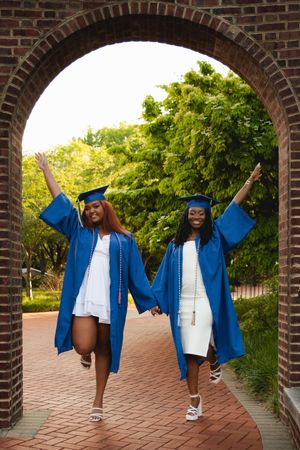 Three graduates in blue caps and gowns jumping joyfully on a brick pathway during celebration photoshoot.