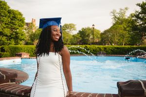 Graduate in blue cap and white dress smiles while standing near a swimming pool on a sunny day.