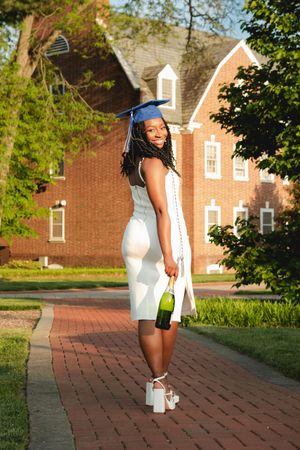 A graduate in a white dress and blue cap poses on a brick pathway with buildings in the background.