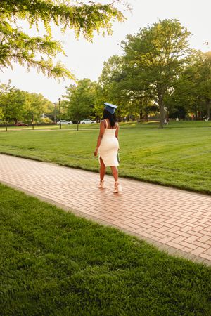 Graduate walks along a winding brick pathway across a sunlit green lawn during golden hour.