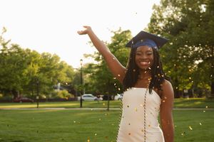 Graduate in cap and gown celebrates with raised arm as confetti falls on sunny green lawn during sunset.