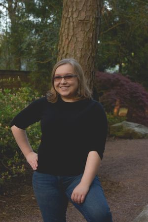 Person in black t-shirt and jeans poses outdoors against backdrop of pine trees and garden landscaping.