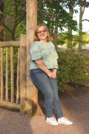 A person in a mint green top and jeans poses casually against a wooden post in an outdoor garden setting.