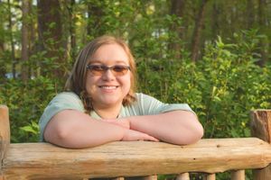 Someone rests their arms on a wooden railing while enjoying a sunny day in a lush green forest setting.