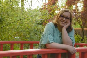 Person in teal shirt leans on red railing in autumn garden setting with trees in background.