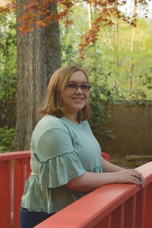 Someone in a mint green blouse leans against a red railing outdoors with autumn foliage in the background.