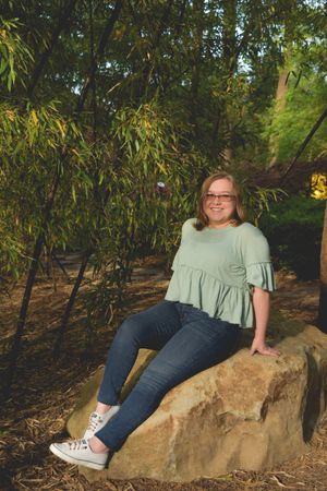 Someone in a mint green ruffled top and jeans sits casually on a large rock surrounded by bamboo plants in a garden setting.