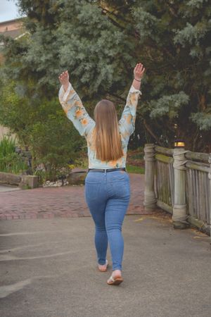 Woman in floral top and blue jeans raises arms joyfully while walking on path with stone railing and trees.