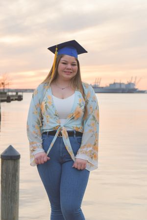 Graduate in blue cap and gown poses at waterfront dock during sunset with harbor and boats in background.