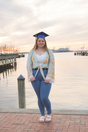 A graduate in blue cap and gown poses on a brick walkway by a waterfront dock during sunset.