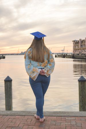 A graduate in blue cap and gown stands on a brick walkway overlooking a waterfront pier at sunset.