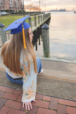 Graduate in blue cap and gown sits on brick walkway by waterfront during sunset.