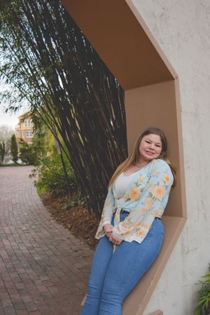 A woman in a floral cardigan and jeans poses in an archway next to bamboo plants.
