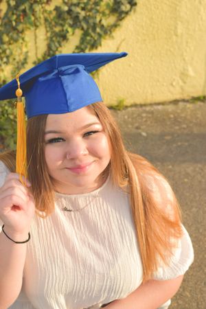Graduate in a blue cap with gold tassel and white dress poses outdoors against yellow wall in sunlight.