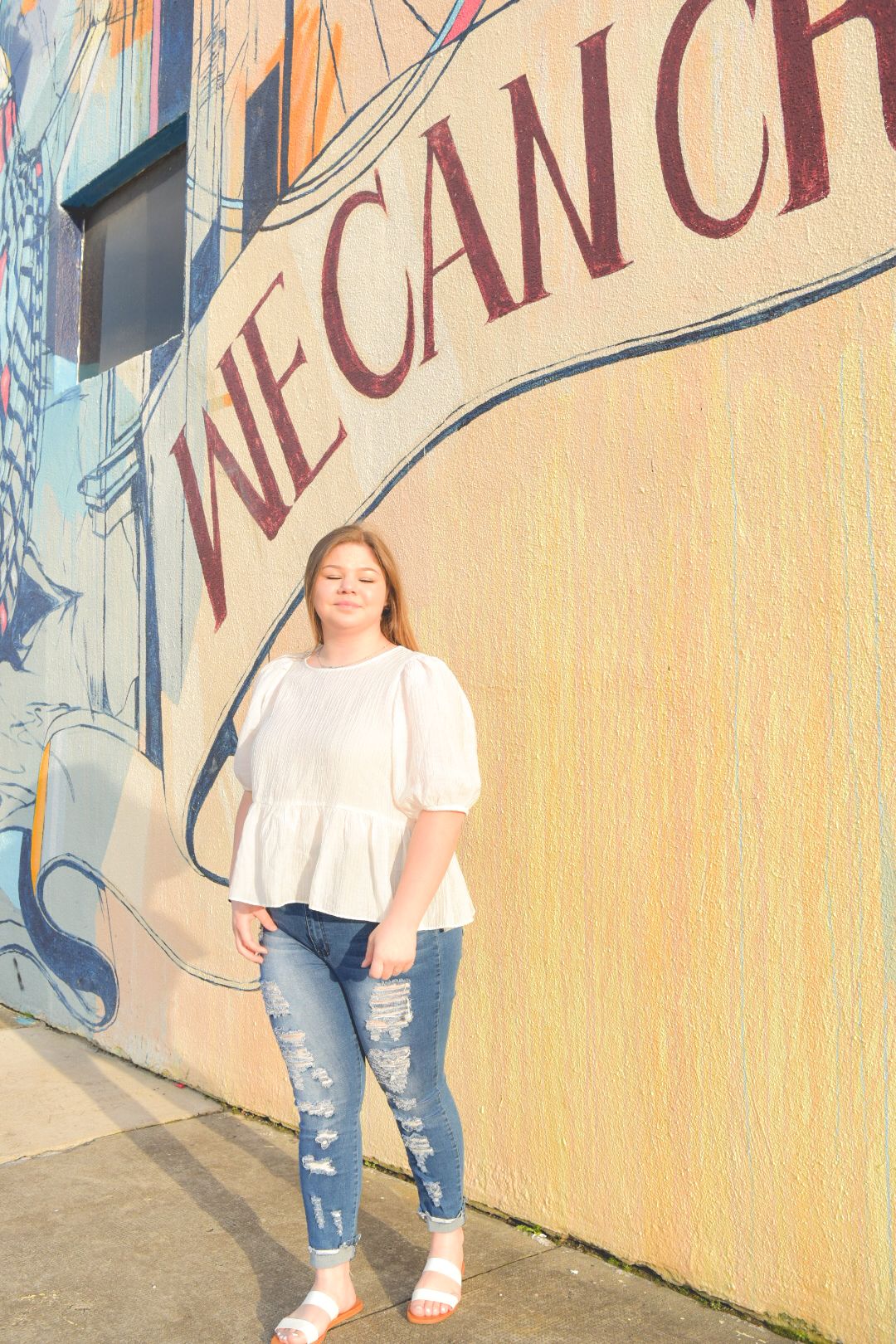 Person in white t-shirt and distressed jeans poses against colorful mural wall with text lettering.