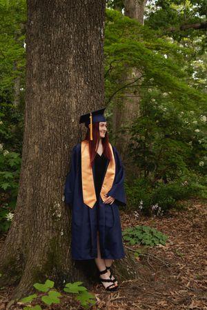 Graduate in navy cap and gown poses near a large tree trunk in a wooded area with orange stole.