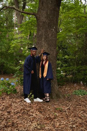 Graduates in navy blue caps and gowns sit together on fallen autumn leaves in a wooded area during a photoshoot.