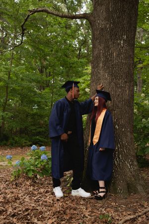 Two graduates in navy blue caps and gowns share a moment by a large tree with blue hydrangeas in the background.
