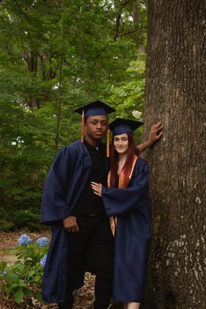 Graduation photos taken in a wooded setting showing graduates in navy blue caps and gowns posing by a tree.