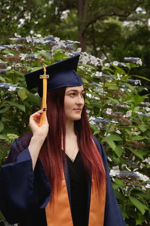 Graduate in navy cap and gown with orange stole poses in front of flowering dogwood trees.