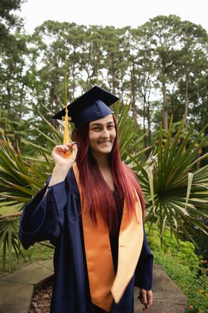 Graduate in navy blue cap and gown with orange stole stands in front of palm trees while making peace sign gesture.