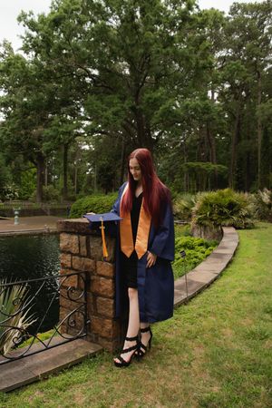 Graduate in cap and gown poses near stone wall and pond in garden setting with curved pathway and lush greenery.