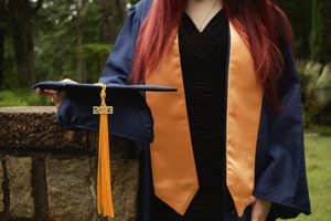 A graduation ceremony detail showing navy blue gowns, orange stoles, and gold tassels against a natural outdoor setting.