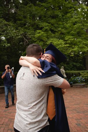 Two people share an emotional embrace on a brick pathway surrounded by green trees in the background.