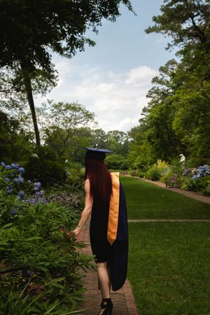 A person in a graduation gown walks along a scenic garden path surrounded by lush green trees and blooming flowers.