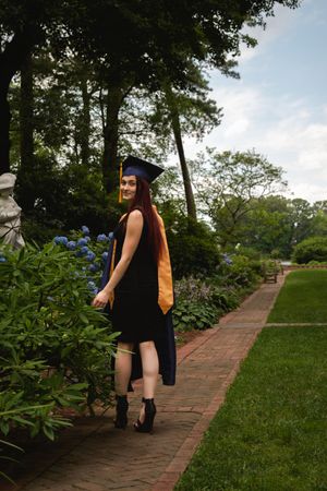 Graduate in cap and gown poses along brick pathway surrounded by lush gardens and hydrangea bushes.