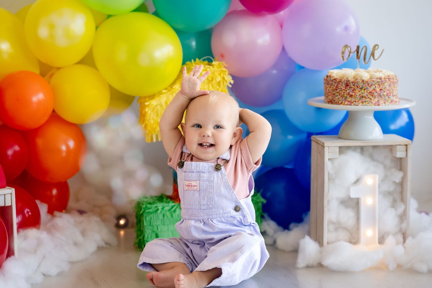Smiling baby in overalls celebrates first birthday with colorful rainbow balloon arch and cake topper reading one.