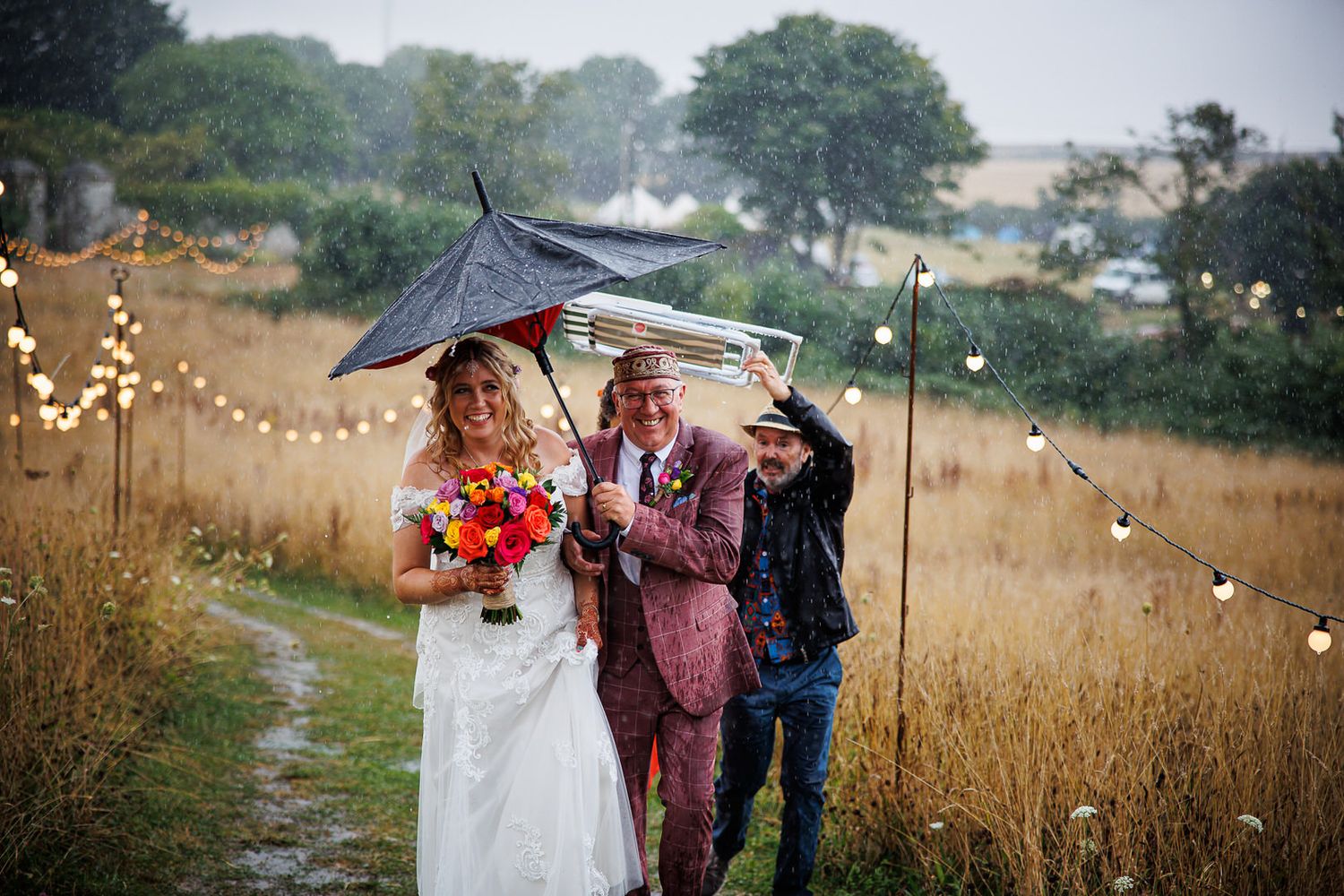 Wedding party walking through a golden field with umbrellas on an overcast rainy day.