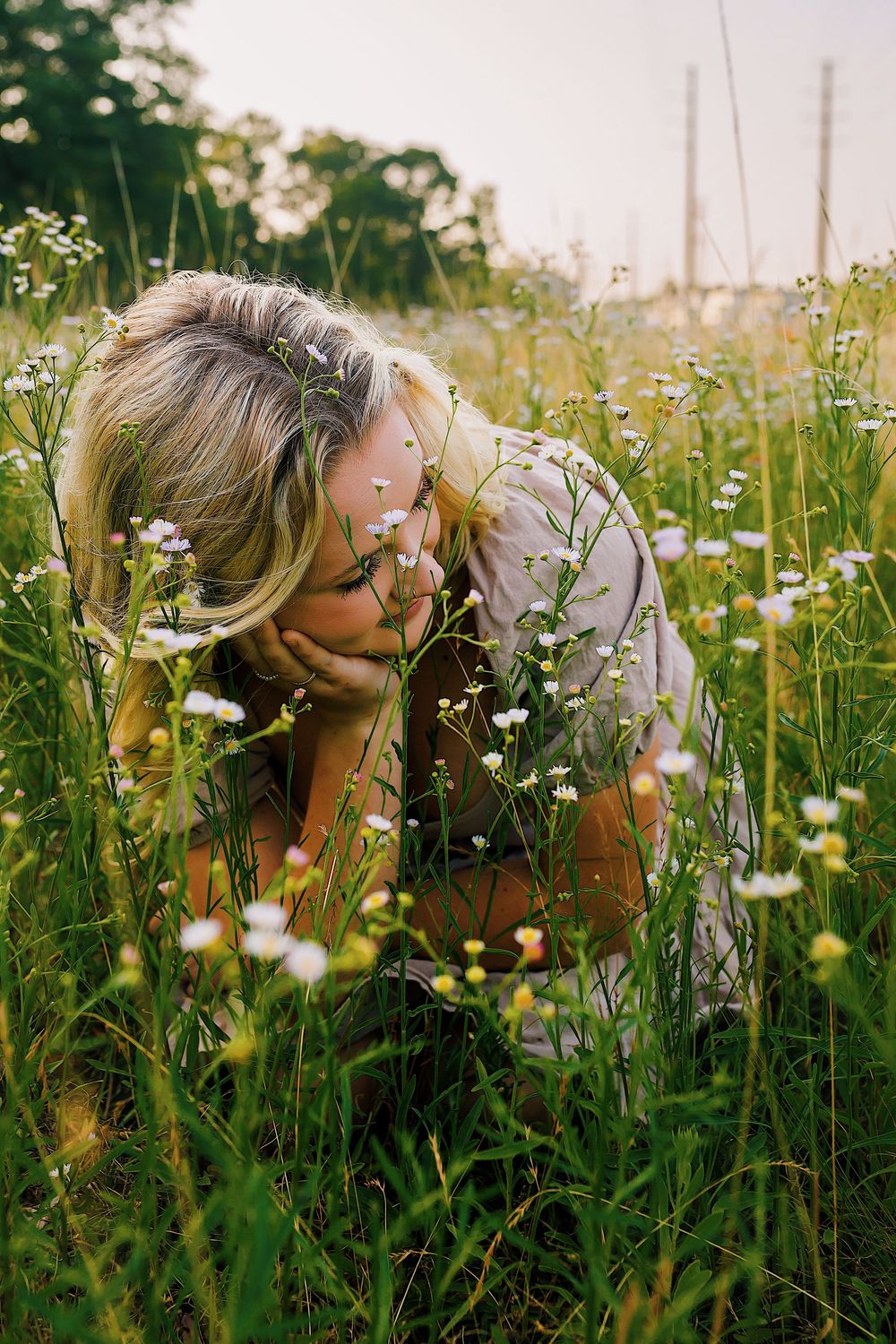 Someone leans down to smell wildflowers in a sunny meadow filled with white daisies and tall grass.