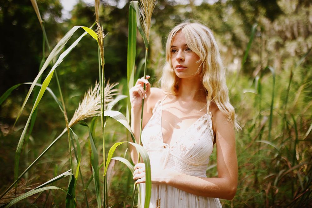 A person in a white lace dress stands among tall grass and wildflowers in a dreamy outdoor setting.