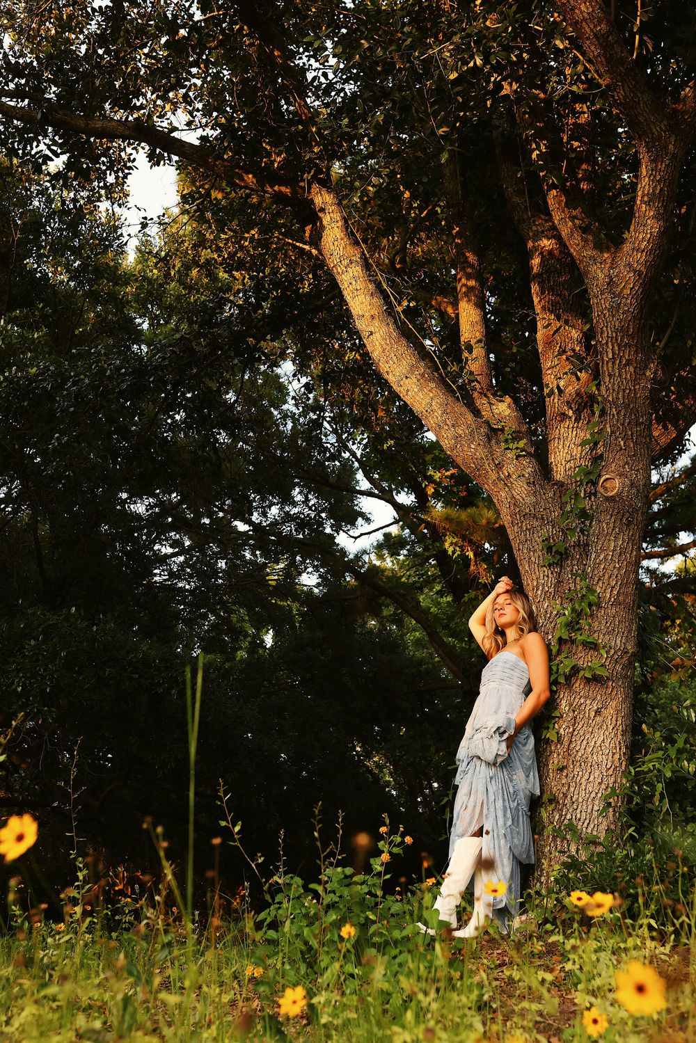 A person in a flowy dress stands beside a large oak tree in a meadow with yellow wildflowers at sunset.