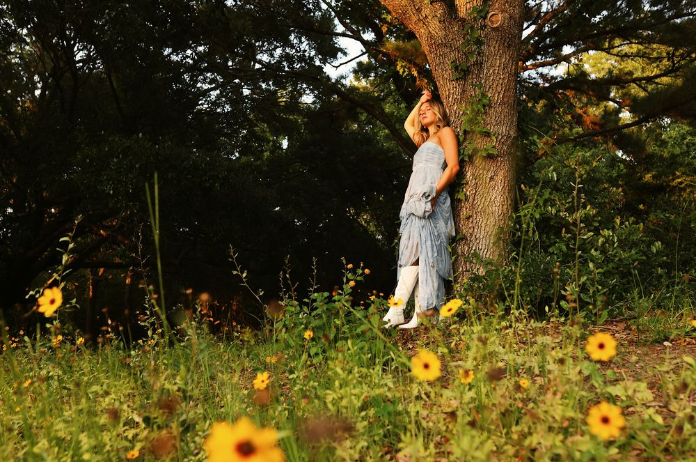 A person in white clothing poses near yellow wildflowers and trees in a scenic outdoor setting during golden hour.