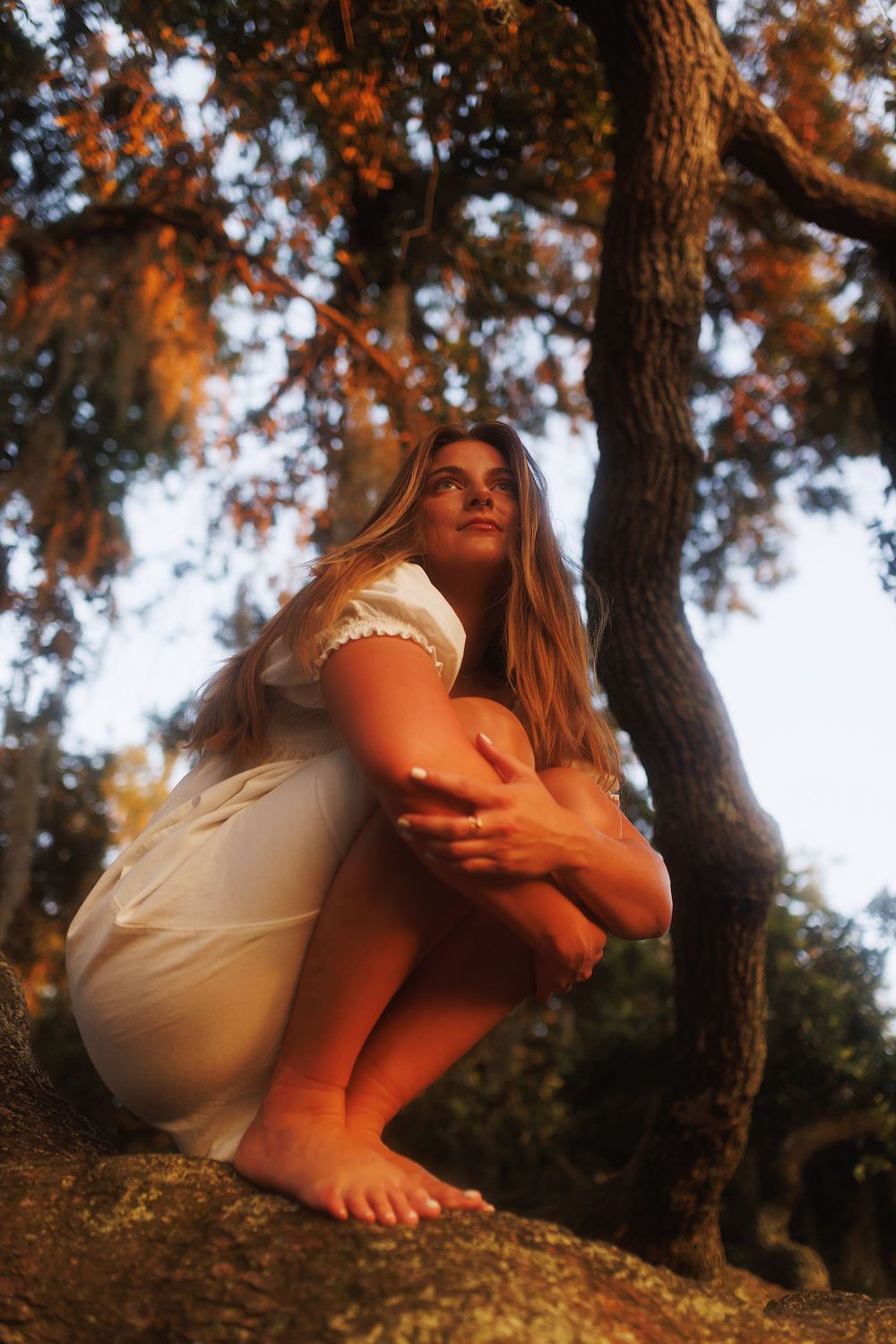 A person in a white dress crouches barefoot on a tree-covered hillside at sunset with warm golden lighting.