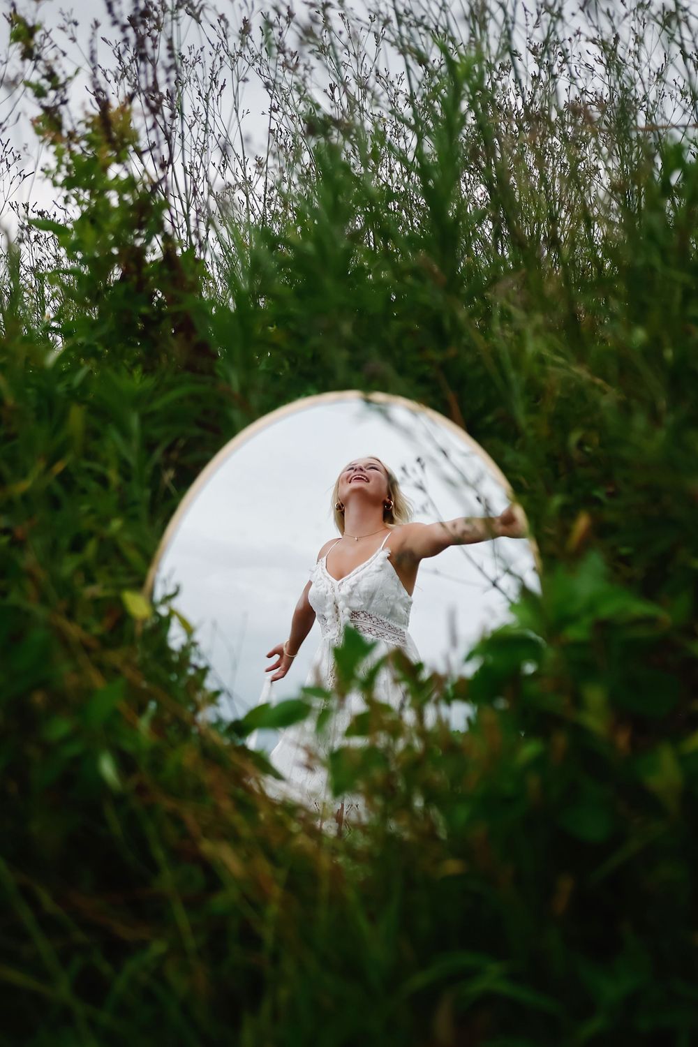 A circular mirror reflects a figure in a white dress against lush green foliage.