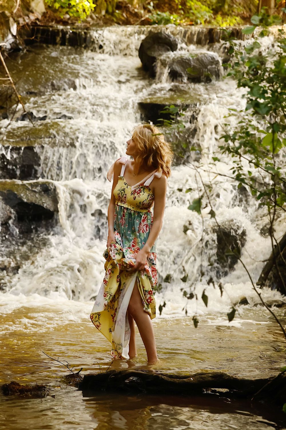 A waterfall cascades over rocks while someone in a floral dress stands near the flowing water on a sunny day.