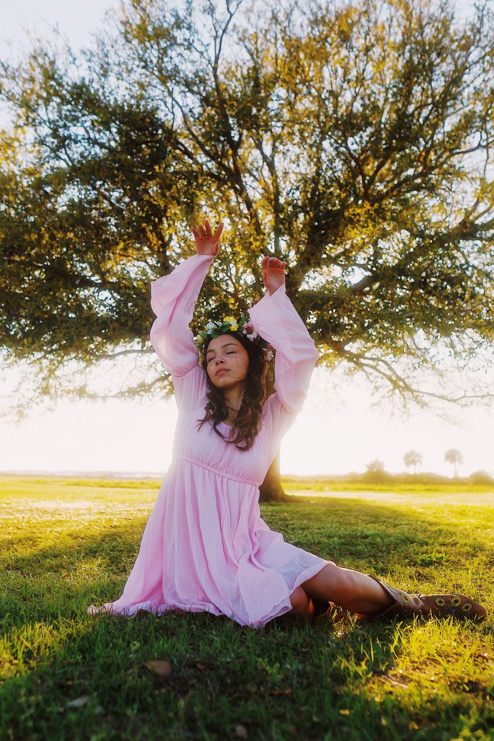 A person in a flowing pink dress sits on grass at sunset, raising their arms joyfully beneath a large tree.