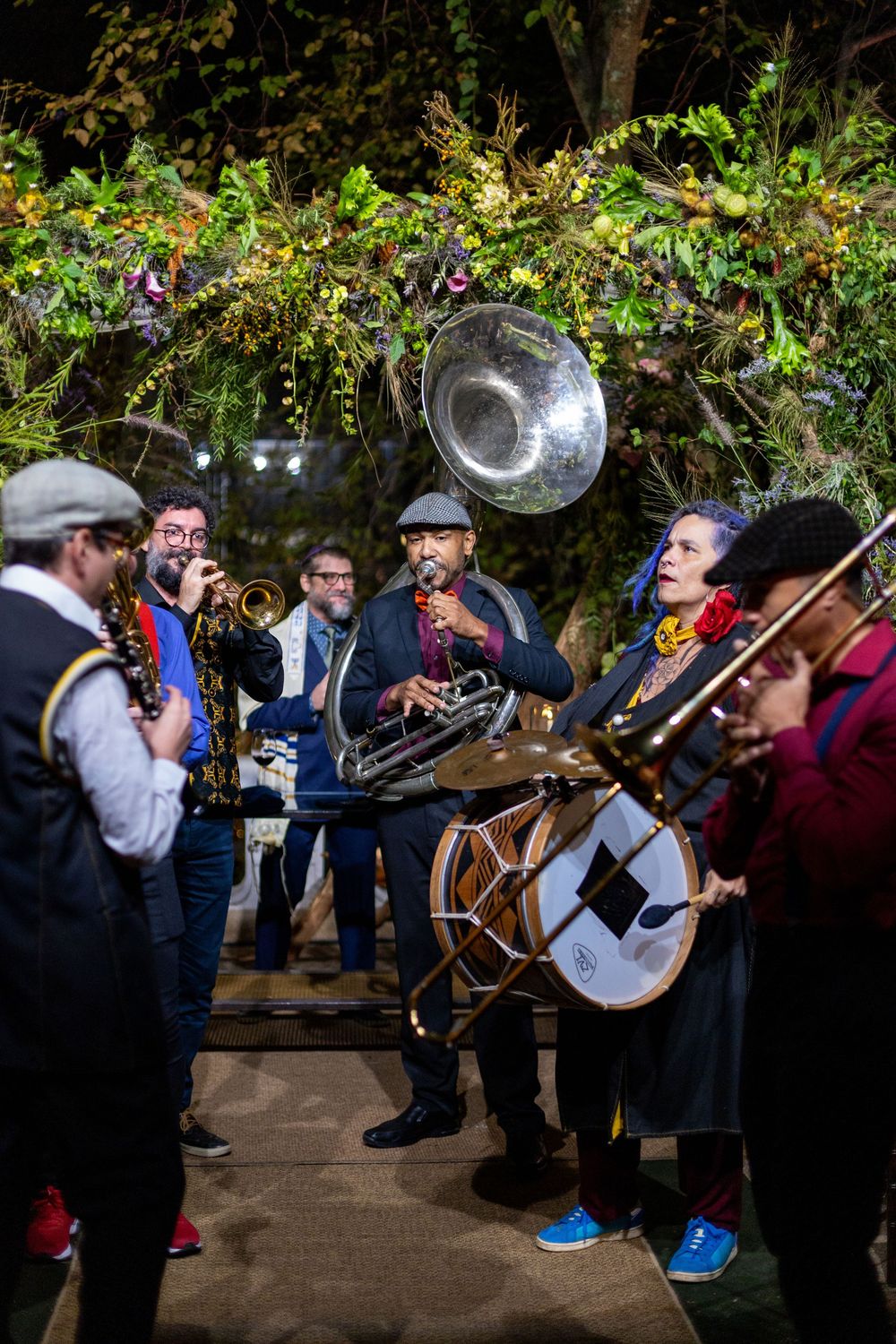 A traditional band with brass and percussion instruments performs outdoors under string lights and foliage.
