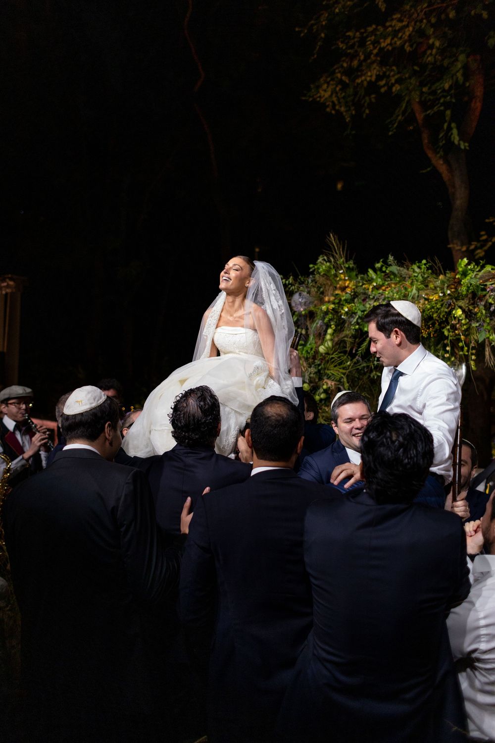 A wedding party in formal attire gathers outdoors with guests wearing dark suits and a white dress visible.
